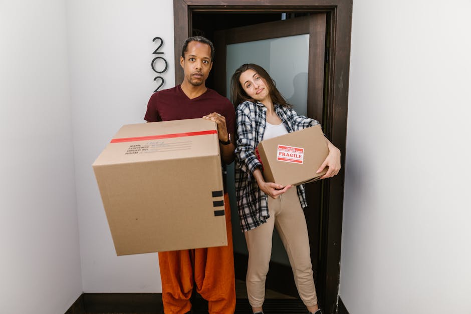 A man and a woman stand inside a doorway, each holding a cardboard box for house relocation. The man, positioned on the left, wears a dark red shirt and orange trousers, carrying a large box with a red stripe and black markings. The woman, on the right, wears a plaid shirt over a white top and beige trousers, holding a smaller box labeled 'FRAGILE' with red and white packaging tape. They are inside a hallway with white walls and a dark wooden doorframe, which appears to be part of an apartment building or residential property; the lighting is neutral. In the background, outside the doorway, a moving trolley or dolly with additional packing materials and boxes can be partially seen, along with a paved outdoor area. The scene depicts the process of packing and preparing for a home relocation, with visible packing materials and an emphasis on furniture transport and moving logistics, as managed by West Kensington Removals.