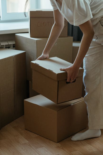 A person wearing a white outfit is seen lifting a cardboard box off a stack of four similar boxes inside a room, with natural light coming through a window behind them. The boxes are plain brown with no visible branding, and one of the boxes on top has 'CLOTH' written on it in red marker. The boxes vary in size and are arranged on a wooden floor, some stacked directly on top of each other, while others are positioned side by side. The scene depicts a home relocation or packing process, supported by the presence of multiple packed boxes indicative of furniture transport and moving preparations, as sometimes managed by professional removals services like West Kensington Removals. The image focuses on the meticulous handling of moving boxes during the packing and home moving process, illustrating the careful loading and transportation of household items.